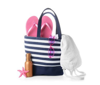 Terlikli çizgili plaj çantası, mayo, güneş gözlüğü, denizyıldızı ve beyaz arka planda bir şişe güneş kremi.