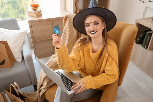 Cadı şapkalı, kredi kartı ve dizüstü bilgisayarı olan genç bir kadın Cadılar Bayramı için internette alışveriş yapıyor.