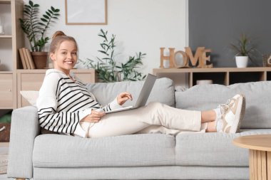 Evdeki koltukta modern laptop kullanan genç bir kadın.