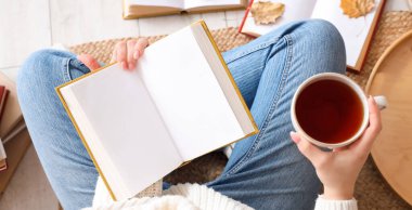 Boş kitabı ve çay bardağıyla evdeki halıda oturan kadın.