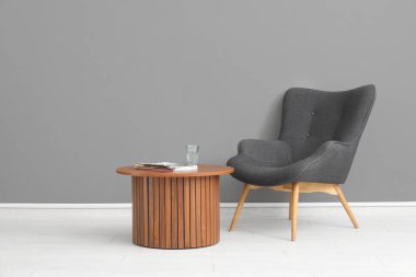 Gri duvarın yanında bir bardak su, dergiler ve koltuk bulunan ahşap sehpa.