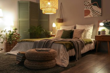 Şık yatak odasının içinde yatağın üzerinde battaniyeler, hasır puflar ve geceleri parlayan lambalar.