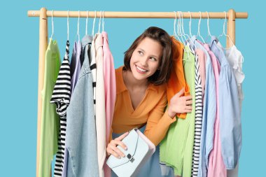 Mavi arka planda farklı giysileri olan, rafların yanında güzel genç bir kadın.