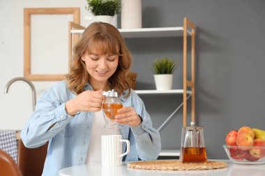 Genç bir kadın mutfaktaki çaya bal ekliyor.