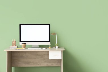 Modern bilgisayarlı şık bir iş yeri, rahat ofisteki yeşil duvarın yanında bir fincan kahve, lamba ve kırtasiye malzemesi