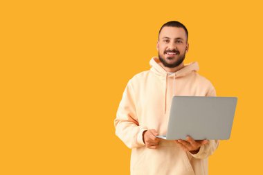 Sarı arka planda dizüstü bilgisayarı olan gülümseyen genç adam.