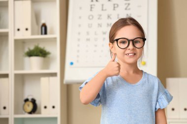 Gözlüklü şirin küçük kız göz doktorunun ofisinde baş parmağını kaldırıyor.