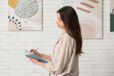 Sanat galerisindeki beyaz tuğlalı duvara, defterin yanına yazı yazan genç bir kadın.