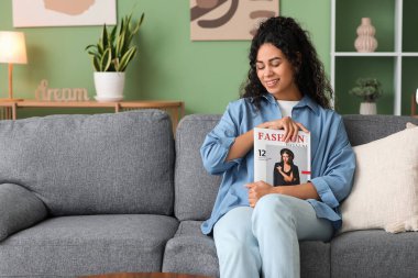 Şık mavi gömlekli, dergisi evdeki kanepede oturan güzel Afro-Amerikan kadın.