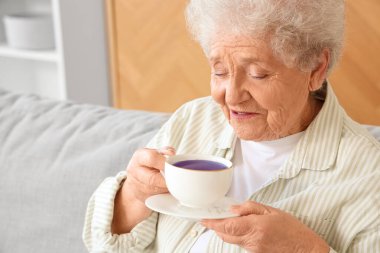 Oturma odasındaki kanepede oturan bir fincan kelebek çiçekli çaylı son sınıf kadını.