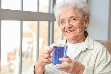 Evinin penceresinin yanındaki cam bardakta bezelye çiçeği çayıyla yaşlı bir kadın.