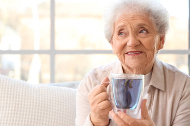 Oturma odasının penceresinin yanındaki cam bardakta bezelye çiçeği çayıyla kıdemli kadın.