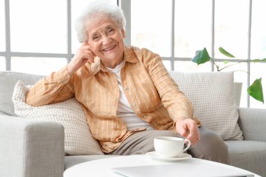 Oturma odasının penceresinin yanındaki kanepede oturan kelebek çiçekli çaylı son sınıf kadını.