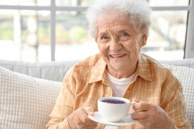 Oturma odasının penceresinin yanındaki kanepede oturan kelebek çiçekli çaylı son sınıf kadını.