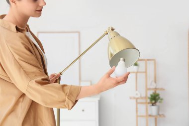 Genç bir kadın evdeki standart lambaya ampul takıyor.