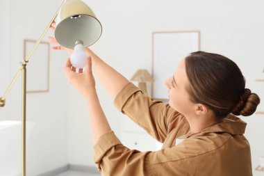 Genç bir kadın evdeki standart lambaya ampul takıyor.