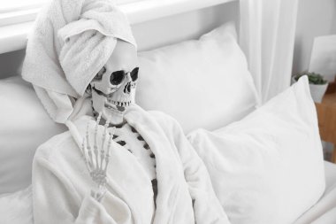 Yatak odasındaki yastıkların üzerinde havlu ve bornozla insan iskeleti modeli.