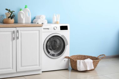 Mavi duvarın yanında sepeti ve dolabı olan modern çamaşır makinesi. Ev çamaşır odasının içi.