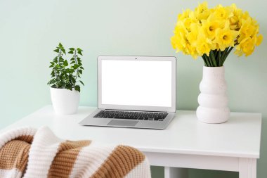 İşyerindeki turkuaz duvarda narsisli çiçekler ve dizüstü bilgisayarı olan bir vazo.