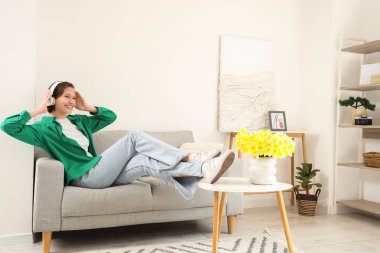 Kulaklıklı genç bir kadın evdeki kanepede müzik dinliyor.