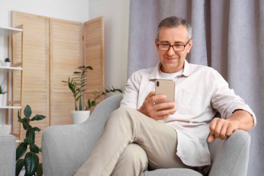 Cep telefonlu yaşlı adam evdeki koltukta oturuyor.