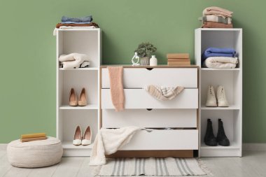 Dağınık oturma odasının içinde raf üniteleri, tuvalet ve dağınık giysiler yeşil duvarın yanında.