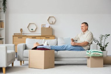 Kanepede dizüstü bilgisayar kullanan yetişkin bir adam. Evde kutular dolusu kıyafet var.