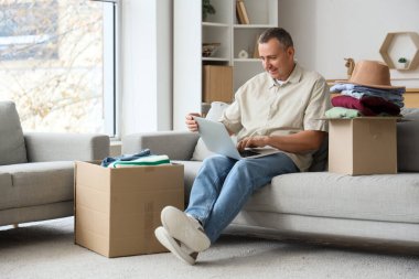 Evde kutular dolusu giysi ve dizüstü bilgisayar kullanan olgun bir adam.