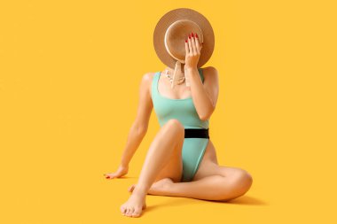 Sarı arka planda oturan hasır şapkalı şık turkuaz mayo giymiş güzel genç bir kadın.