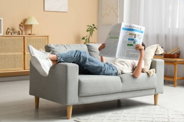 Evdeki gri koltukta gazete okuyan genç bir kadın.