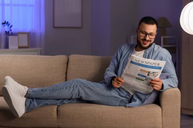 Evdeki kanepede gözlüklü yakışıklı bir adam gazete okuyor.