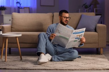 Gözlüklü yakışıklı adam, akşam evde halıda gazete okuyor.