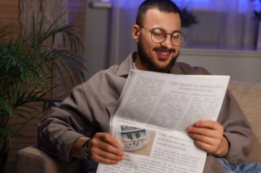 Evdeki kanepede gözlüklü yakışıklı bir adam gazete okuyor.