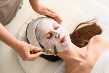 Masaj maskesiyle kozmetik uzmanı güzellik salonunda kadının yüzüne maske takıyor.