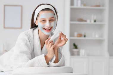 Kil maskeli ve kozmetik ürünü şişesiyle kaplıca salonundaki kanepede uzanan güzel, genç ve mutlu bir kadın.