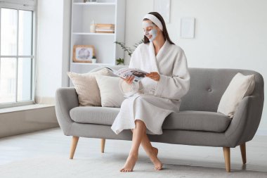 Evdeki kanepede kilden maske okuyan bornozlu, genç ve güzel bir kadın.