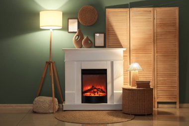 Gece oturma odasının içinde katlanan ekran, parlayan lambalar, hasır masa ve mavi duvarın yanındaki şömine dekoru.