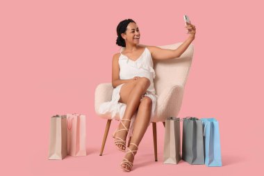 Güzel, genç, mutlu, Afro-Amerikan bir kadın, alışveriş torbaları ve cep telefonuyla pembe arka planda selfie çekiyor.