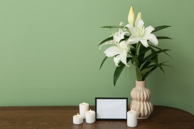 Boş cenaze çerçevesi, yanan mumlar ve vazo, ahşap dolapta, renkli duvarın yanında zambak çiçekleri.