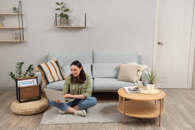 Evdeki gri kanepenin yanında dizüstü bilgisayar kullanan genç bir kadın.