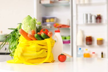 Mutfak tezgahında açık buzdolabının yanında içinde ürünler ve şişe olan sarı alışveriş torbası.