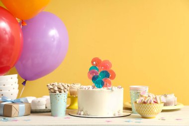 Sarı duvara karşı doğum günü partisi için içinde pasta, şeker ve balon olan bir masa.