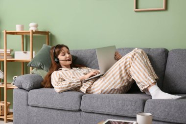 Pijamalı ve kulaklıklı genç bir kadın dizüstü bilgisayarla çalışıyor ve evdeki kanepede yatıyor. Uzak çalışma konsepti