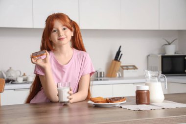 Masaya yaslanmış tatlı bir kız. Yanında bir kavanoz çikolata ezmesi, süt ve mutfakta tatlı tost var.