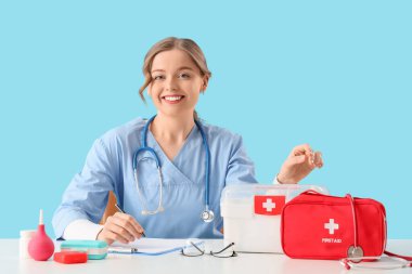 Mavi arka planda, masada oturan, ilk yardım çantası ve panosu olan kadın doktor.