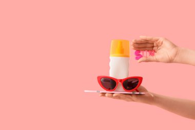 Güneş gözlüklü ve pembe arka planda güneş kremi şişesi tutan kadın.