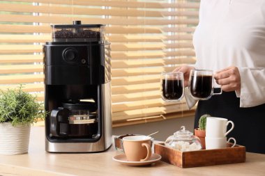 Mutfaktaki kahve makinesinin yanında elinde kahve fincanı olan genç bir kadın.