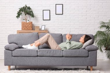 Genç Asyalı kadın evdeki yumuşak koltukta cep telefonu kullanıyor.