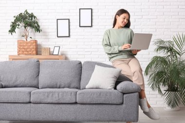 Genç Asyalı kadın evdeki yumuşak koltukta dizüstü bilgisayar kullanıyor.