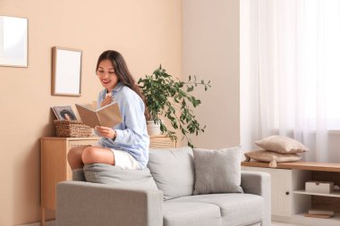 Genç Asyalı kadın evdeki gri koltukta kitap okuyor.